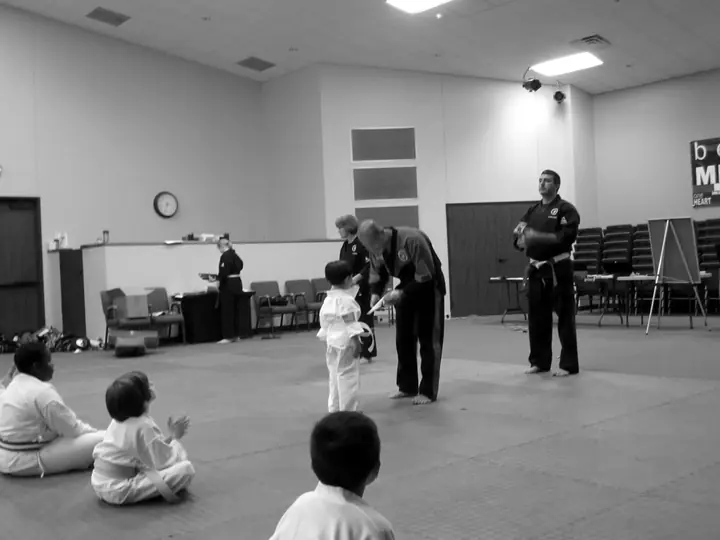 Instructor Y.J. Kim tying a new belt on a young student while classmates watch